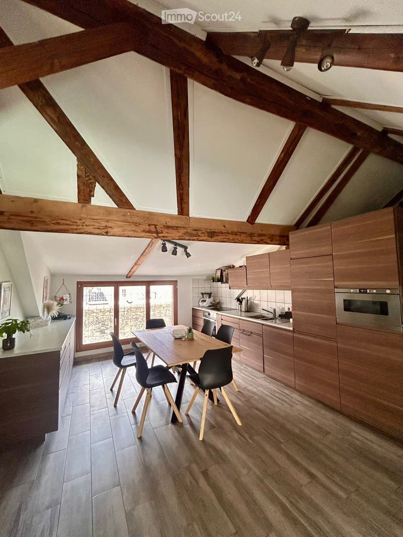 Modern kitchen with wooden beams, a dining table with chairs, wooden cabinets, stainless steel appliances, and a large window overlooking a stone wall.