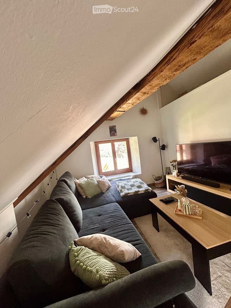 Cozy attic living room with slanted wooden ceiling beams, grey sectional sofa, coffee table, TV, and large window with wooden frame.