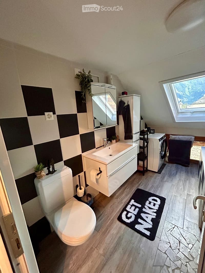 Modern bathroom with a toilet, sink, mirror, cabinet, washing machine, and a black rug with white text.