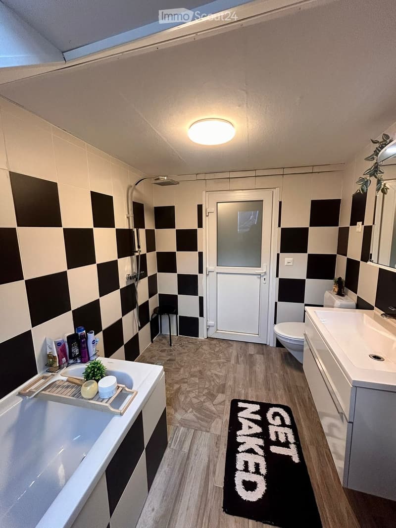 Modern bathroom with checkered black and white tiles, wooden flooring, bathtub, sink, toilet, shower, and a small stool. Includes a black bath mat with white text and a small plant on the sink.