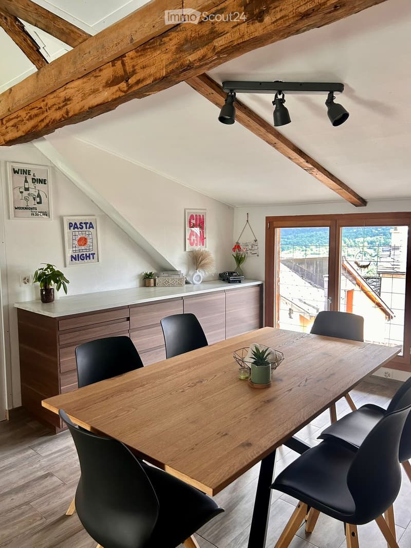 Modern dining area with wooden table, black chairs, exposed wooden beams, and a view through large windows.