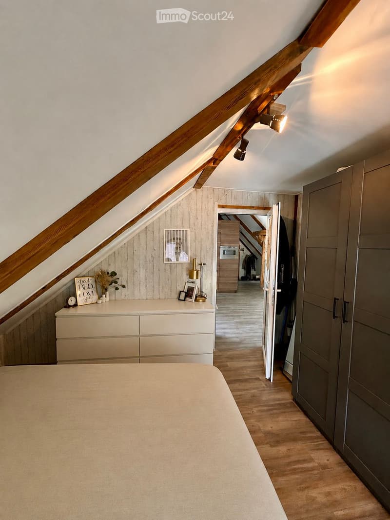 Cozy attic bedroom with wooden slanted ceilings, a large bed, a white dresser with decorative items, and an open doorway leading to another room.