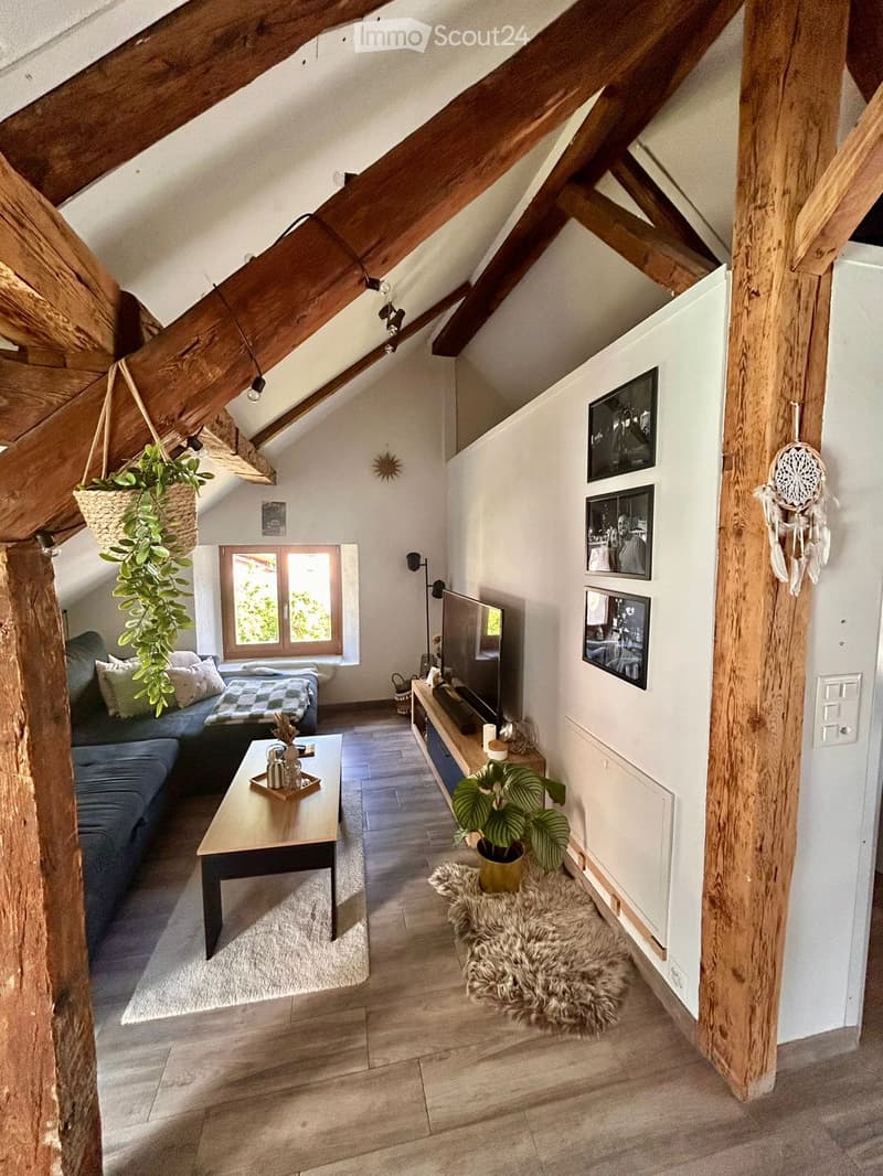 Cozy attic living space with wooden beams, exposed wooden ceiling, large window, sofa, coffee table, TV, and decorative plants.
