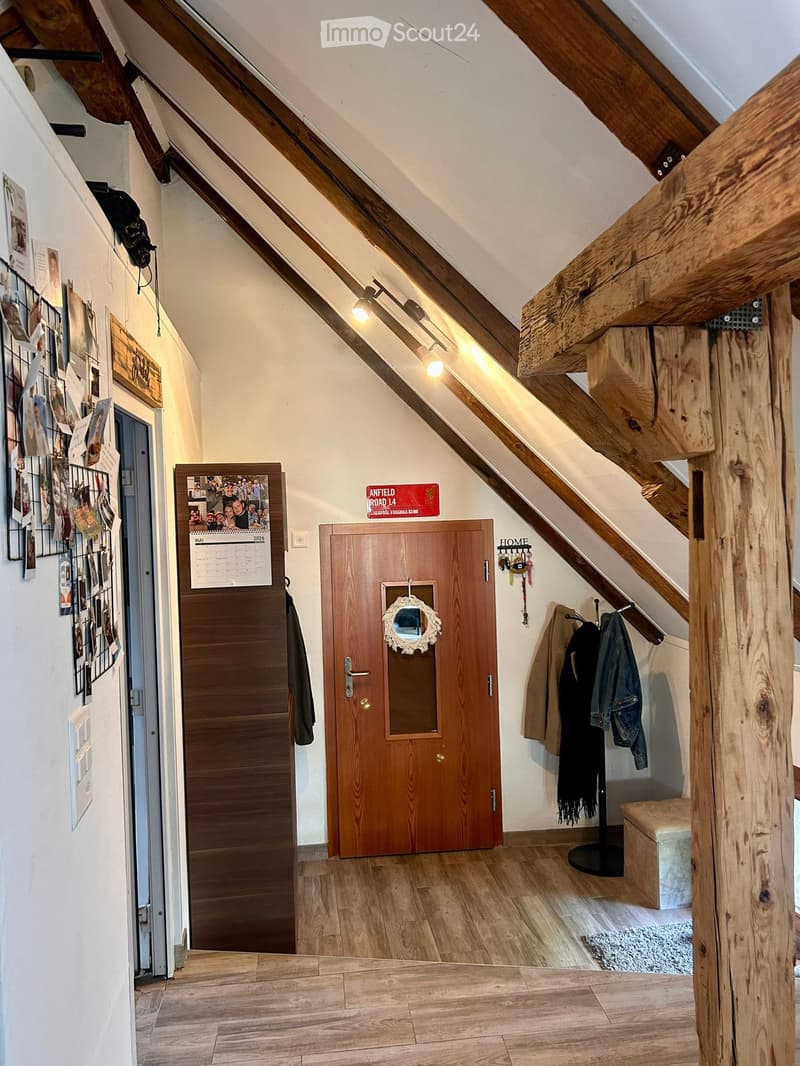 Slanted wooden ceiling hallway with exposed beams, wooden floor, coat rack, calendar, and a small bench.