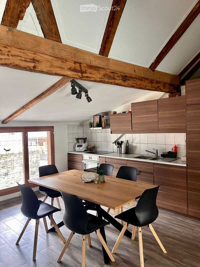 Modern kitchen with wooden cabinets, a large wooden dining table with six black chairs, and various appliances including an oven, sink, and blender. The space features exposed wooden beams and a skylight.