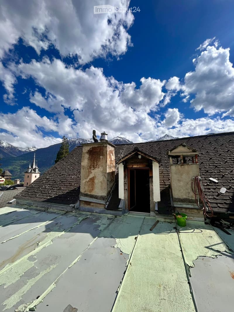 Roof of a building with a chimney, small entrance, and distant mountain view under a partly cloudy sky.