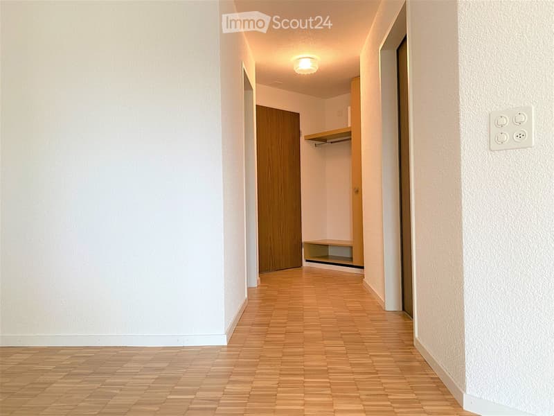 A clean, well-lit hallway with wooden flooring, white walls, and a built-in wooden shelf unit. A closed wooden door is visible on the left, and a light fixture is mounted on the ceiling.