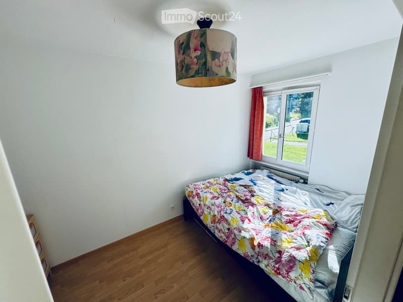 Bedroom with wooden flooring, a single bed with a colorful floral duvet, a hanging lamp with a patterned shade, and a window with red curtains.