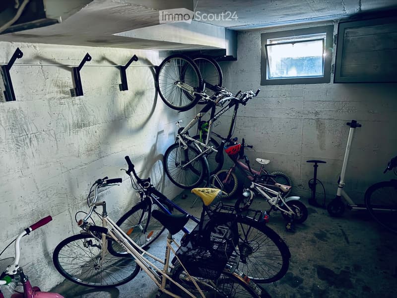 Underground parking area with several bicycles and a scooter stored, concrete walls, and a small window letting in natural light.