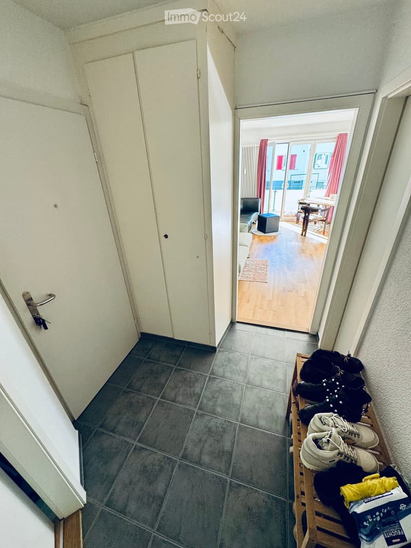 A narrow hallway with tiled flooring, a closed door on the left, and an open doorway leading to a room with wooden flooring and furniture. A small wooden shelf holds shoes and other items.