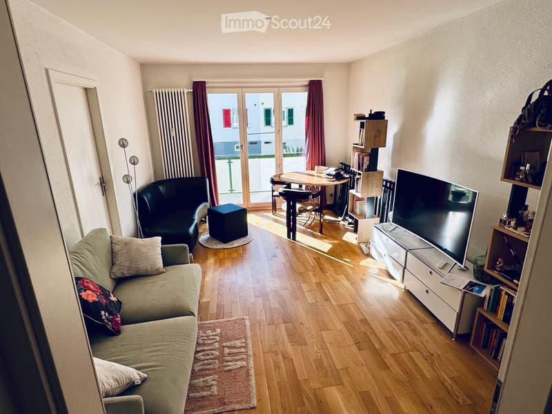Modern living room with light wooden flooring, a grey sofa with floral cushions, a black ottoman, a small black armchair, a coffee table, a TV stand, and a bookshelf.