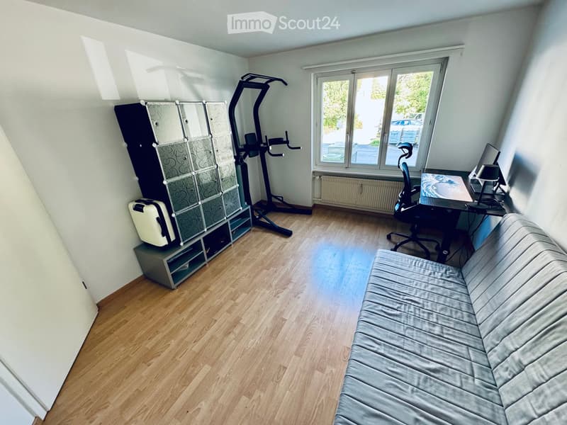 A multi-functional room with a sofa, desk, chair, lamp, storage unit, and a small fitness machine. The room has wooden flooring and large windows letting in natural light.
