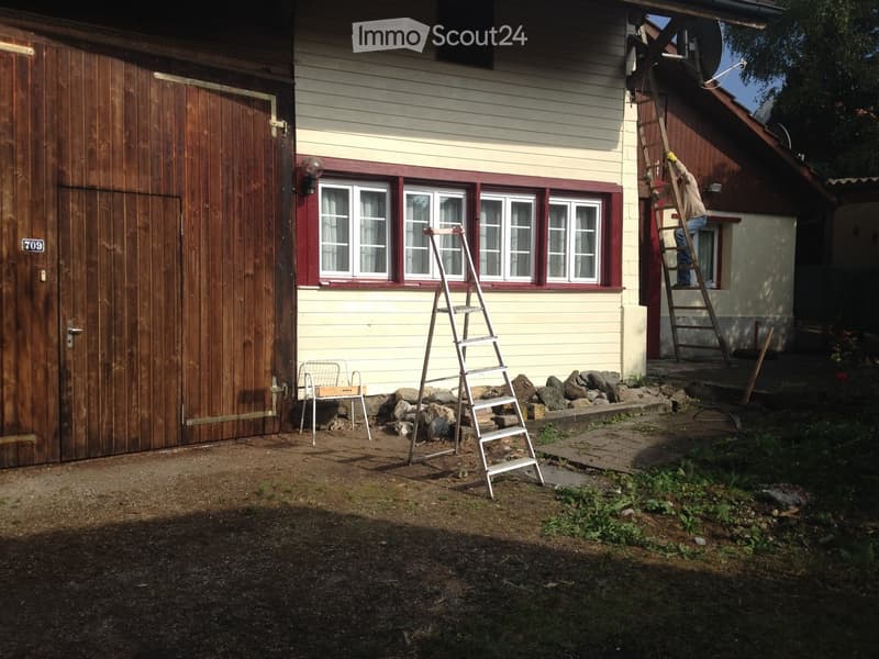A residential outdoor area with a house, garage, ladders, and a small chair. The house has light yellow siding, red trim around the windows, and a wooden garage door. There are two ladders leaning against the house, a small white metal chair, and some roc