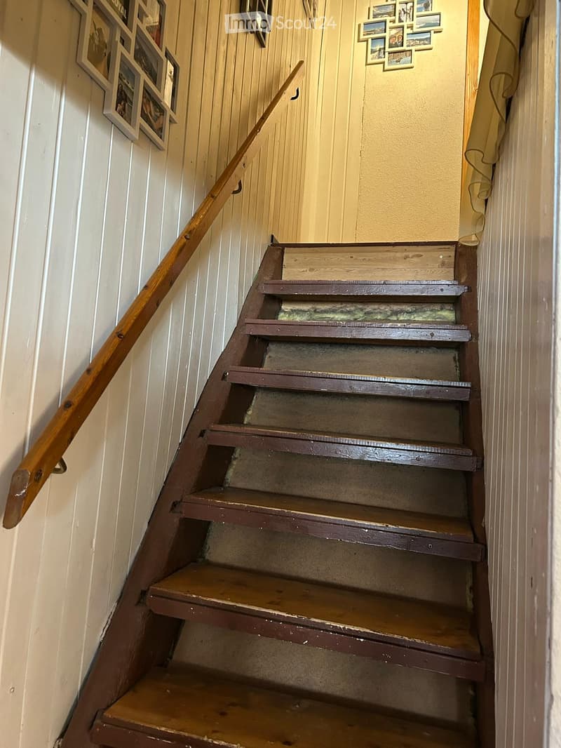 Wooden staircase with a handrail, featuring a mix of wooden and painted steps, flanked by paneled walls and framed pictures.