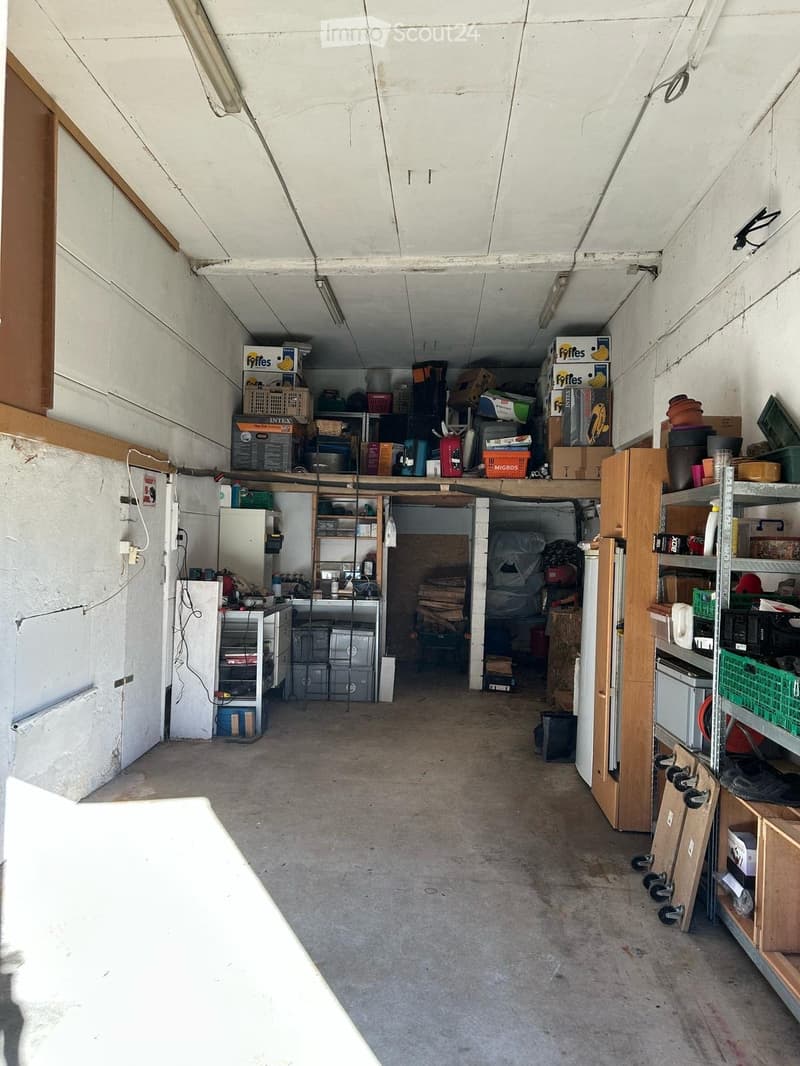 A spacious, unfinished garage with concrete flooring, white walls, and exposed wooden beams. Shelves and storage bins line the walls, with a refrigerator and worktop visible on the left side.