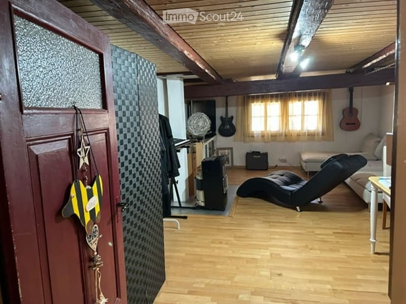 A cozy, wooden-floored room with exposed wooden beams on the ceiling. Features include a black leather sofa, a white armchair, a guitar hanging on the wall, a small table with a lamp, and a cabinet with a speaker system.