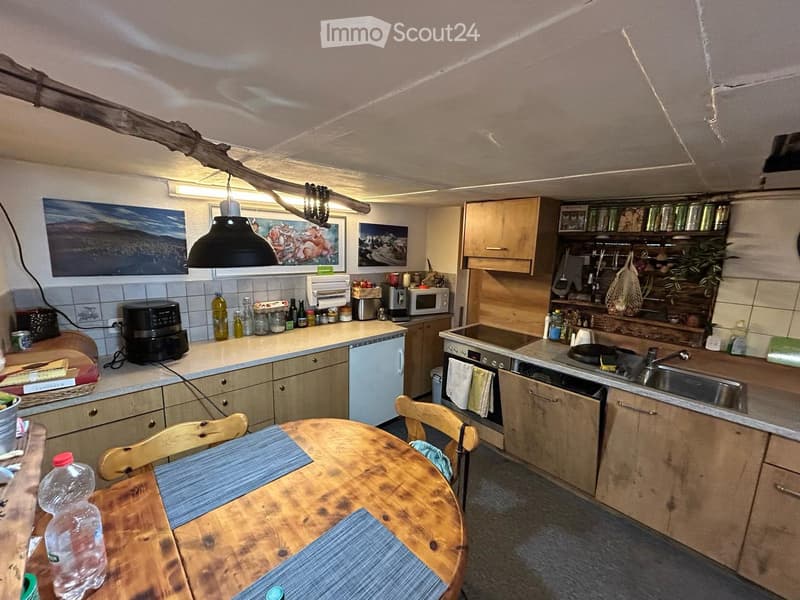 The kitchen features wooden cabinets, a central island with a sink, a dining table with chairs, and various appliances including a microwave, oven, and air fryer. There are also decorative elements like artwork and a hanging light fixture.
