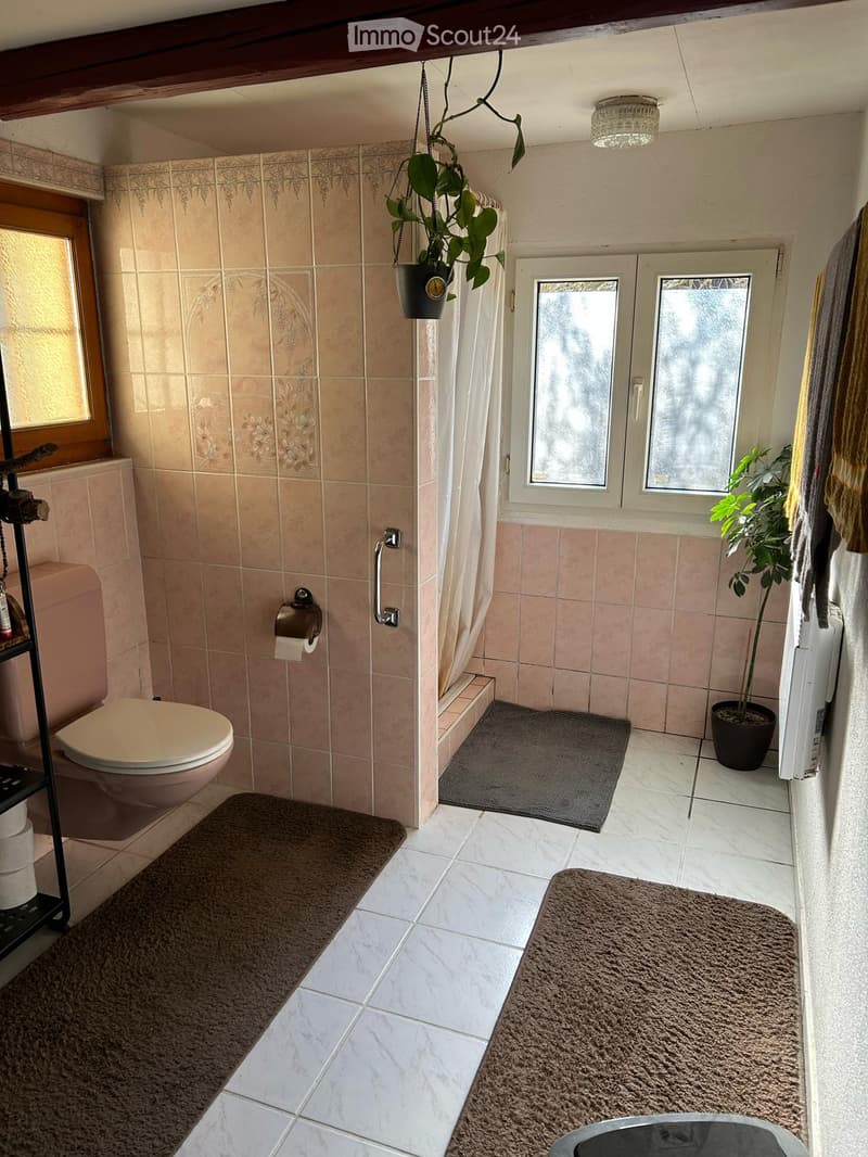 Bathroom with beige tiles, toilet, shower area with curtain, potted plants, and rugs on the floor.