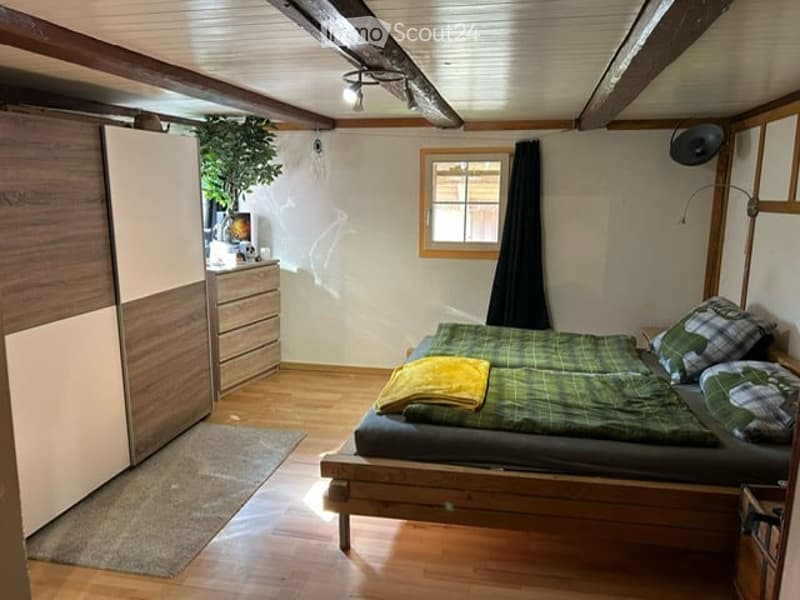 Cozy bedroom with wooden bed, green plaid bedding, wardrobe, nightstand, lamp, and small plant. Wooden floor and slanted ceiling beams.