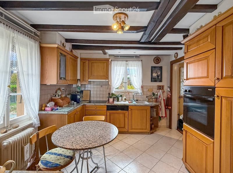 The kitchen features wooden cabinets, a granite countertop, a sink, and a built-in oven. There is a round table with a chair, a window with curtains, and a radiator near the window.