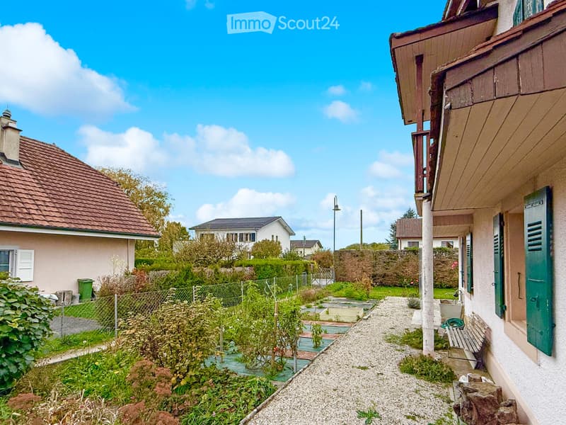 A narrow garden path leads to a house with a wooden bench and green shutters. The garden is enclosed by a fence and contains various plants and shrubs.