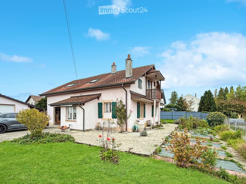 Two-story house with a tiled roof, green shutters, and a balcony. The front yard features a well-maintained lawn, garden beds, and a small patio area with outdoor furniture.