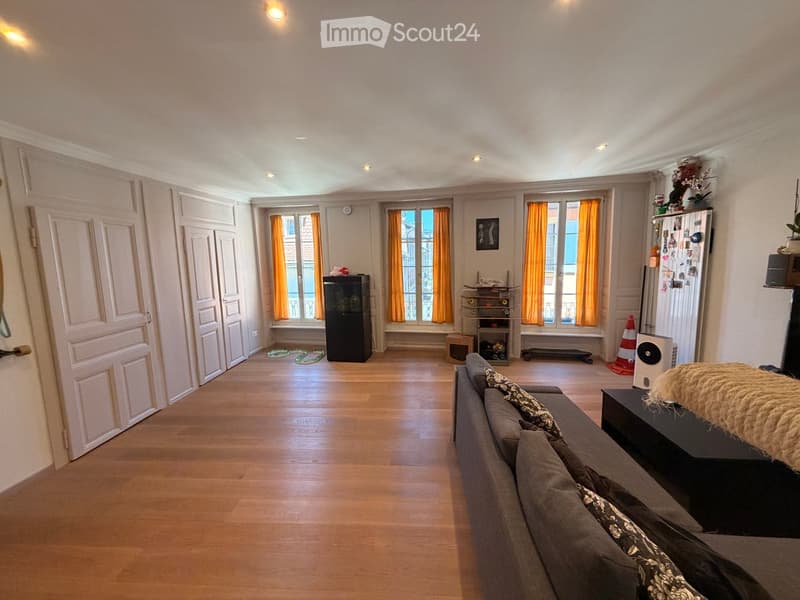 A spacious living room with wooden flooring, large windows with orange curtains, a grey sofa with patterned cushions, a black table, a refrigerator, and a cabinet. The room also features a skateboard and a speaker on the floor.