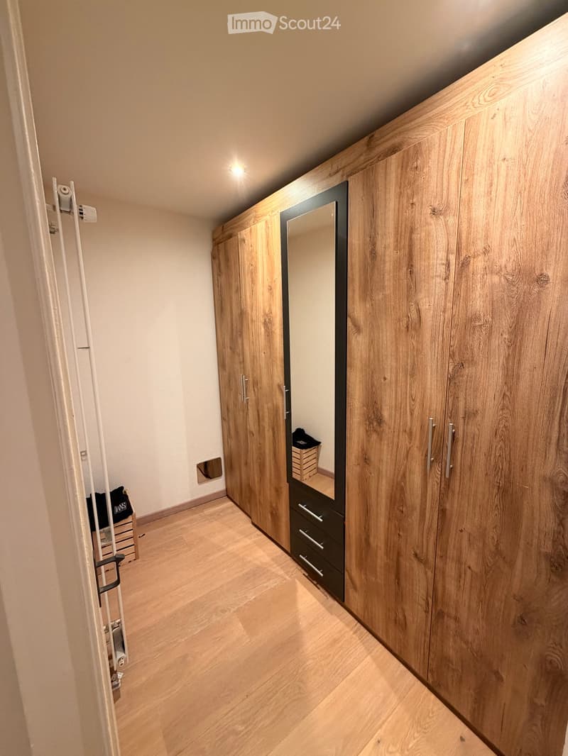 A narrow hallway with light wooden flooring and white walls, featuring built-in wooden wardrobes with mirrored doors and drawers.