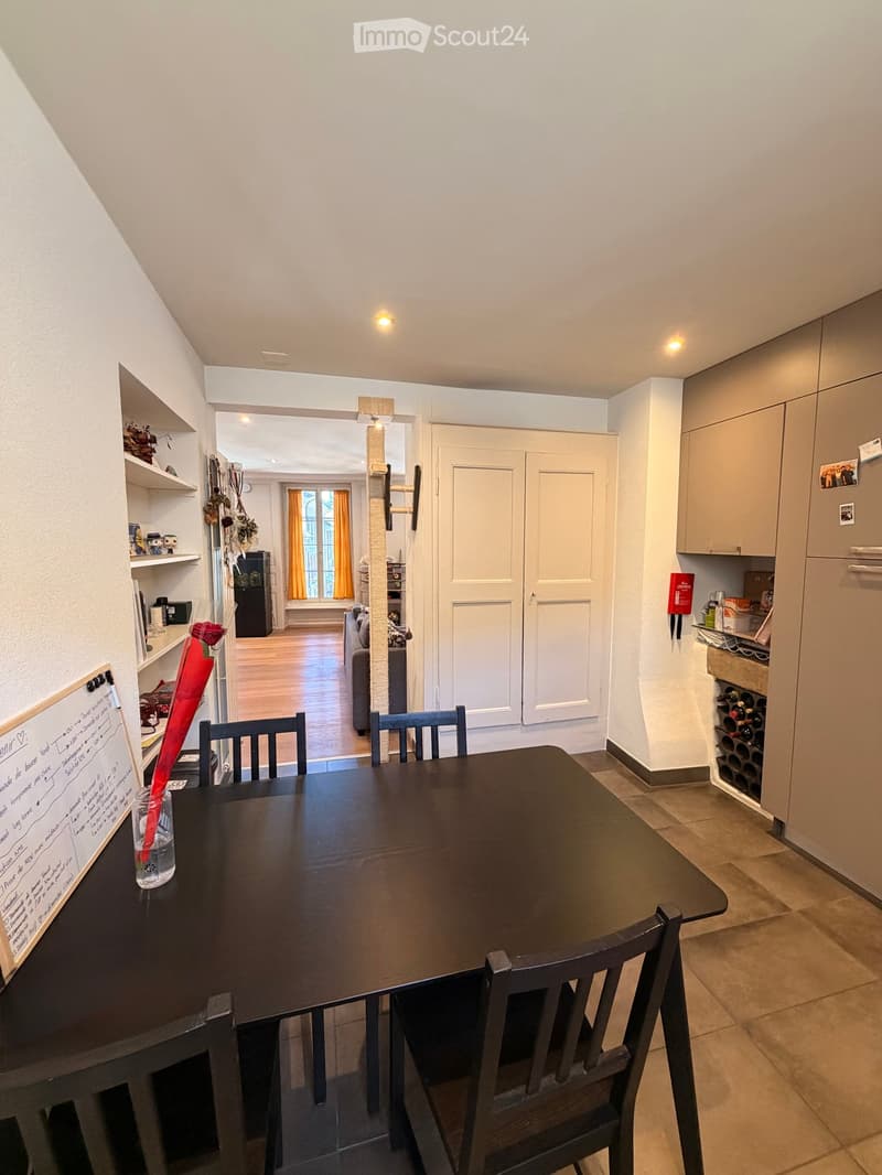 Dining area with a dark wooden table and chairs, a cat tree in the background, shelves with various items, and a kitchen area with cabinets and a wine rack.