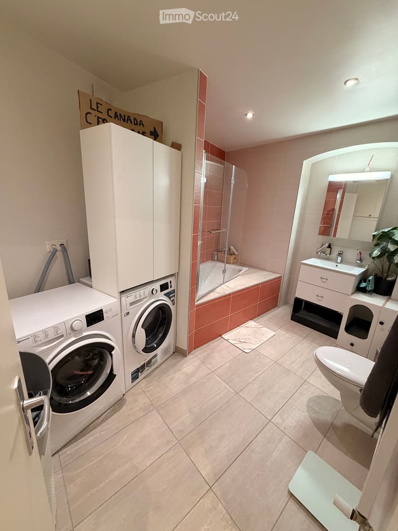 Modern bathroom with a washing machine and dryer unit, a bathtub with a glass shower enclosure, a sink with a large mirror, and a toilet. The space features light-colored tiles and a small plant on the sink counter.
