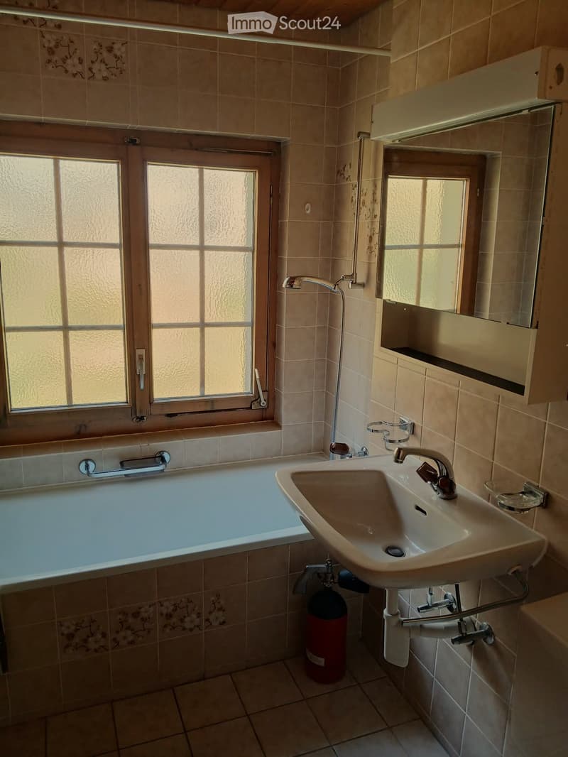 Bathroom with beige tiled walls and floor, featuring a bathtub, sink, mirror, and cabinet. A window with frosted glass panes allows natural light to enter.