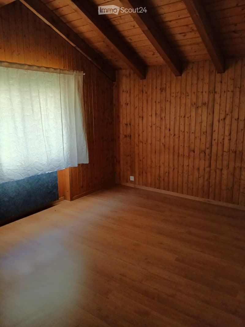 Empty attic room with wooden walls and ceiling, featuring exposed wooden beams and a single window with white curtains.