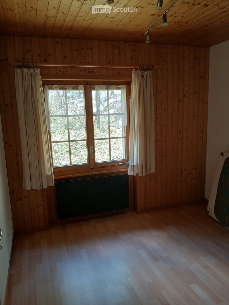 Empty room with wooden walls, wooden floor, large windows with curtains, and a radiator below the windows.