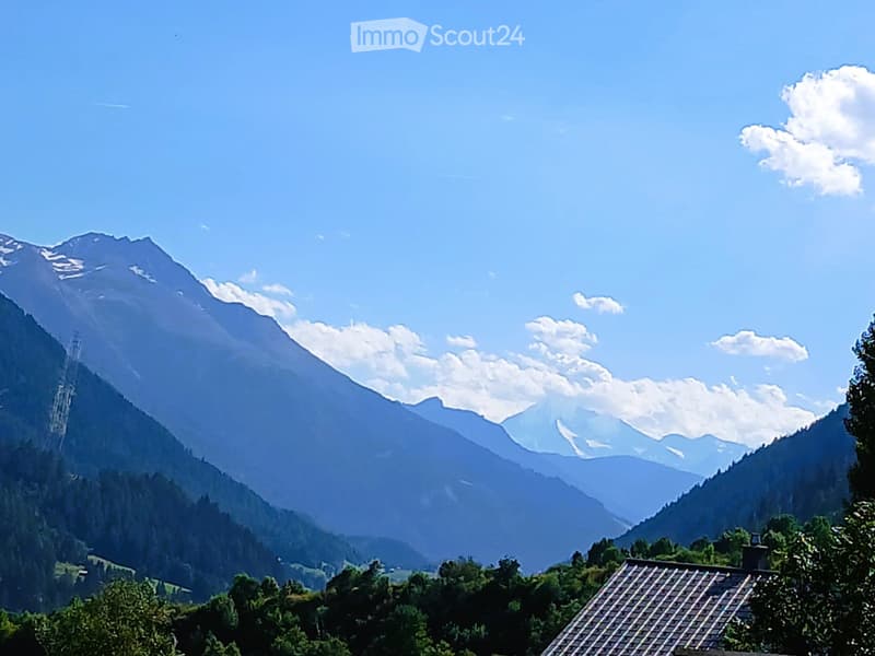 A scenic mountain landscape with a clear blue sky, snow-capped peaks, and a small house partially visible in the foreground.