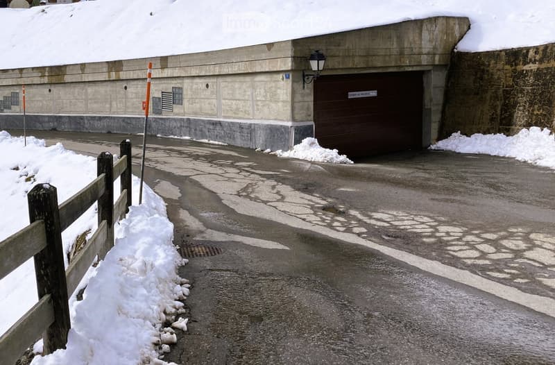 A concrete building with a large garage door, snow on the roof and ground, and a wooden fence along the sidewalk.