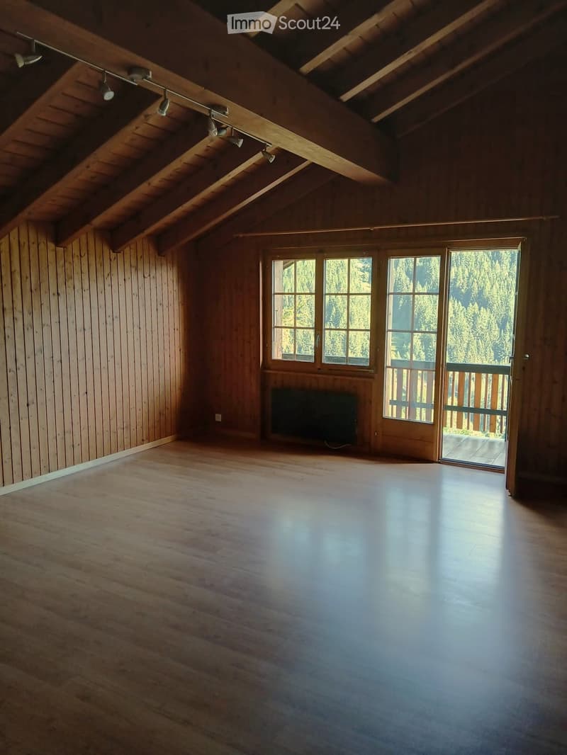 Empty wooden room with exposed beams, large windows, and a sliding door leading to a balcony.