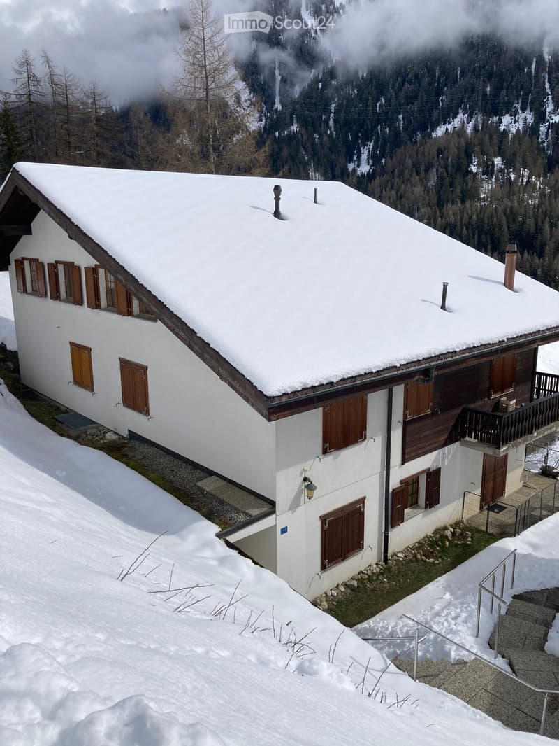 A two-story white house with a snow-covered roof and wooden shutters, situated in a snowy mountainous area with trees and a view of the surrounding landscape.