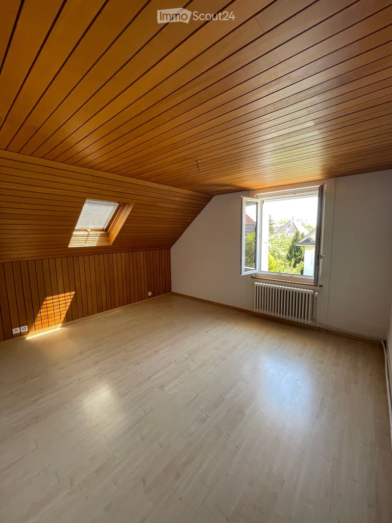 Empty attic room with wooden walls, ceiling, and floor, featuring two windows and a radiator.