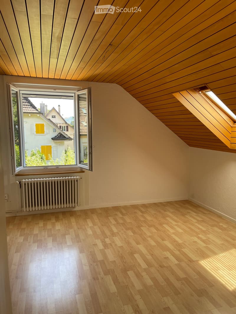 Empty attic room with wooden flooring, slanted wooden ceiling, large window with view of neighboring houses, and a radiator below the window.