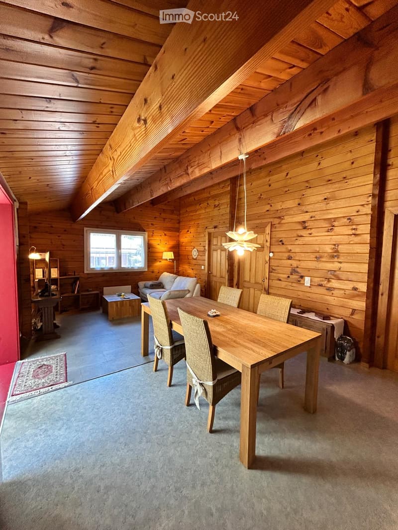 Cozy, wooden-paneled room with a dining table, chairs, sofa, and a lamp. The room features exposed wooden beams on the ceiling and a large window letting in natural light.