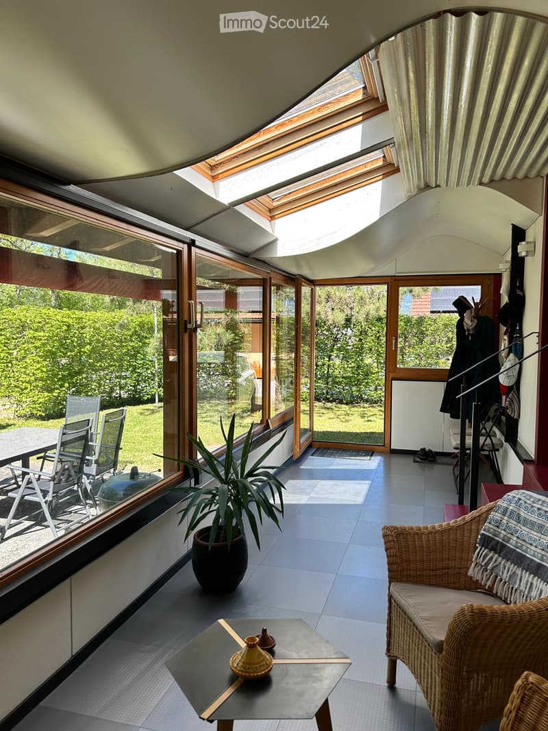 Bright, modern living space with large windows and a skylight, featuring a wicker chair, a small table with a potted plant, and a coffee table with decorative items.
