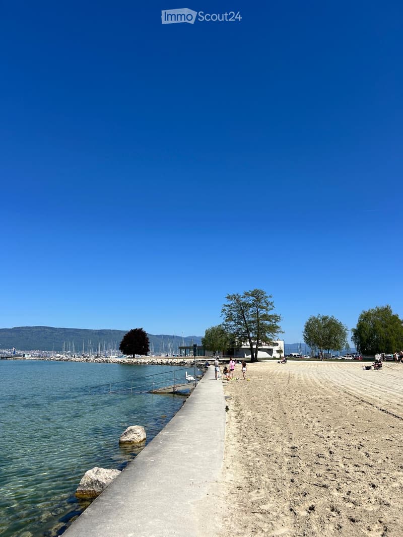 A sandy beach with a concrete walkway, calm water, and distant mountains. A few people are visible near the water's edge.