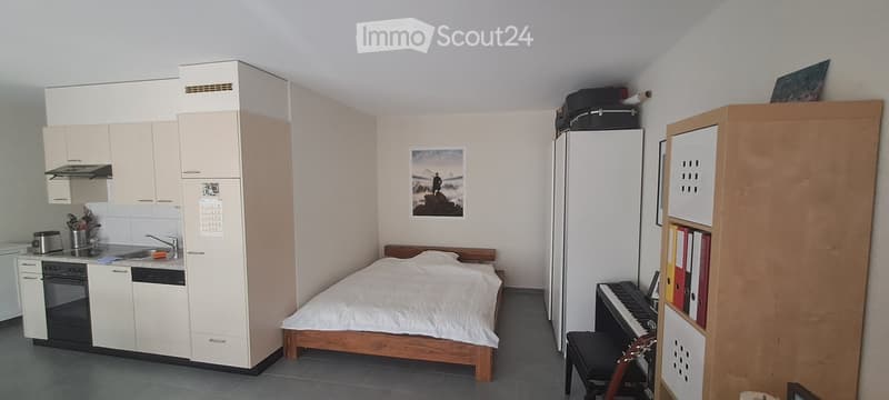 A compact studio apartment with a bed, small kitchen area, refrigerator, piano, and bookshelf. The kitchen features white cabinets, a sink, and an oven. A picture frame hangs on the wall above the bed.