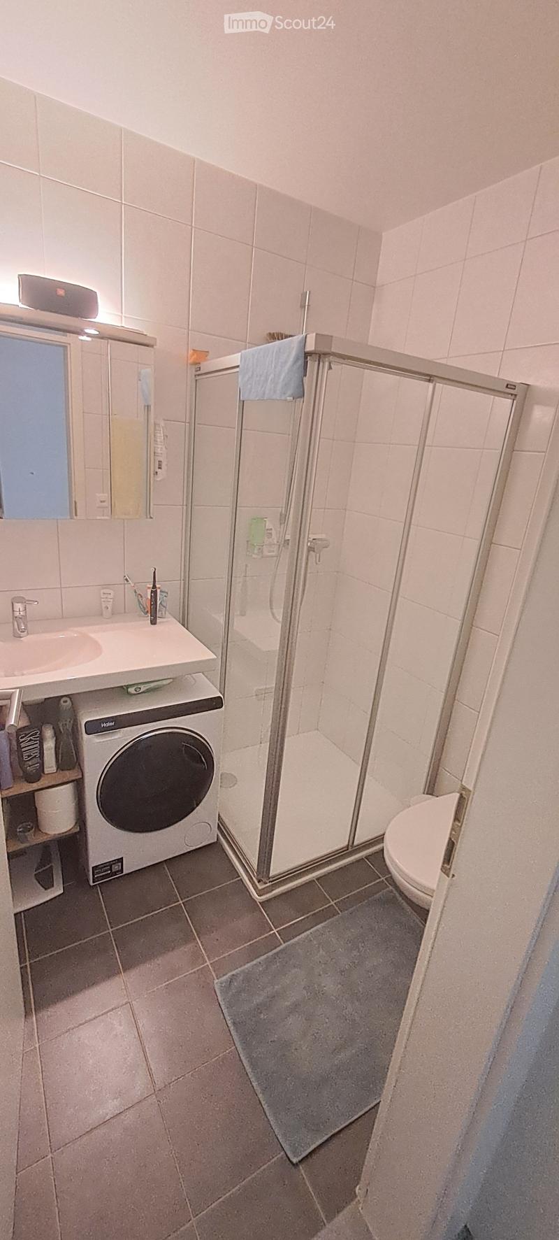 Modern bathroom with white tiles, a sink with a mirror, a shower with a glass door, a toilet, and a washing machine. A towel hangs on the shower door, and a bathmat is placed on the floor.
