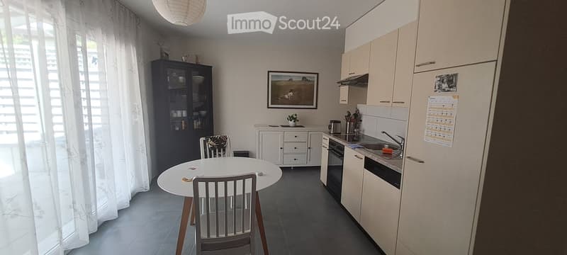 Modern kitchen with light-colored cabinets, a white dining table with chairs, a sink, and a refrigerator. The space is bright with large windows covered by white curtains and includes a pendant light fixture.