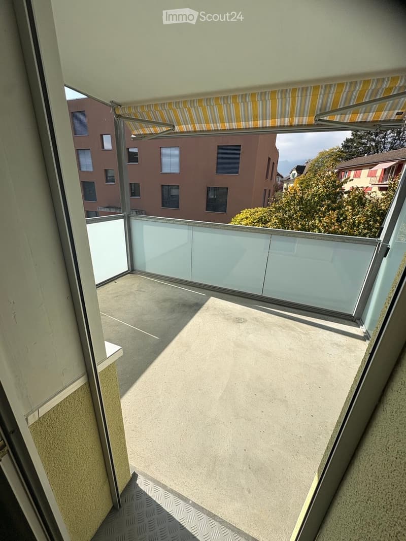 Balcony with glass railing, concrete floor, and striped awning, overlooking neighboring buildings and greenery.