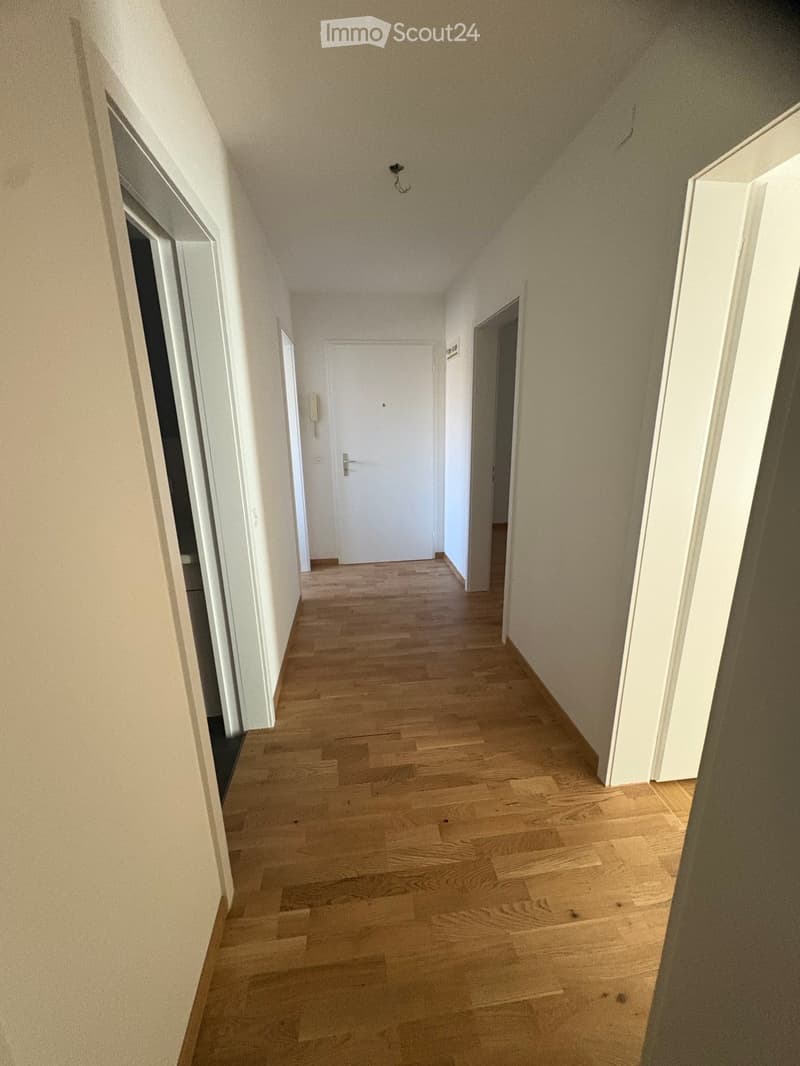 A clean, well-lit hallway with wooden flooring and white walls, featuring several doors and a visible light fixture on the ceiling.