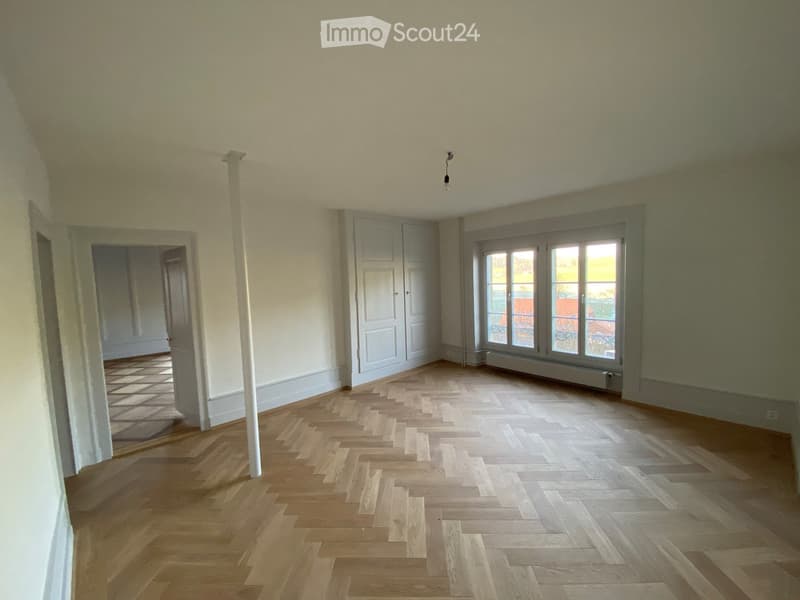 Empty room with wooden herringbone flooring, white walls, and large windows with a view of the outside. There is a white column and a door leading to another room.