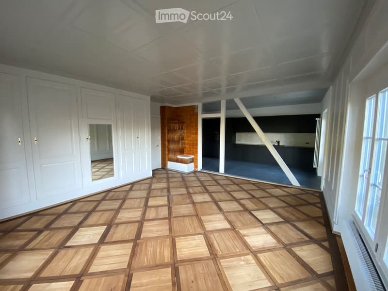 Empty room with wooden parquet flooring, white walls, large windows, and a visible kitchen area in the background.