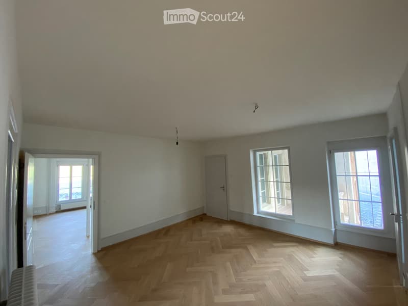 Empty room with wooden herringbone flooring, white walls, and large windows allowing natural light. The room features a door leading to another space and a radiator on the left side.